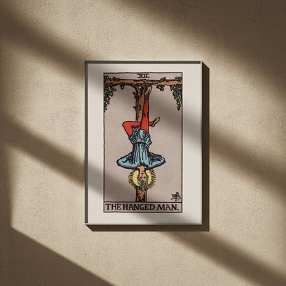Le Pendu, arcane XII du tarot Rider-Waite en 50x70, encadré sur un mur beige, ombres de fenêtre